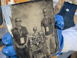 Gloved hands hold an angled black-and-white glass plate portrait of two people flanking a robot.