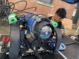 Blue cylindrical underwater robot with a front camera and multiple thrusters rests on a worktable.