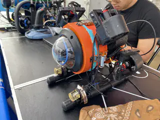 Orange-and-blue underwater robot displays its domed front camera, side thrusters, and exposed wiring.