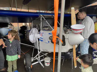 Children gather around a small propeller-driven aircraft with a blue wing beneath a canopy.
