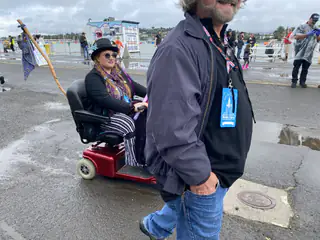 Person riding a red mobility scooter passes behind another attendee on wet pavement.