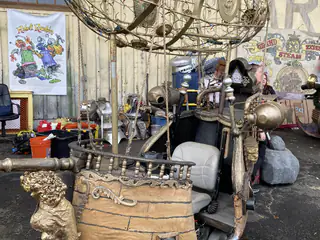 Gold boat-shaped steampunk vehicle sits beneath an oval wire balloon decorated with metal parts.