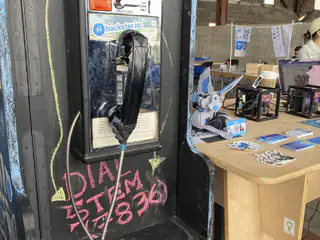 Black wall-mounted telephone handset hangs below a Hackster sticker and pink chalk writing.