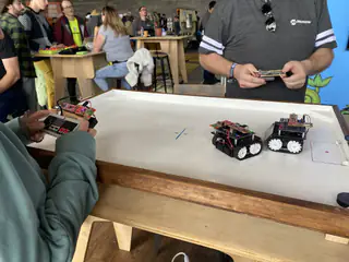 Two people operate handheld controllers while small wheeled robots face each other on a tabletop.