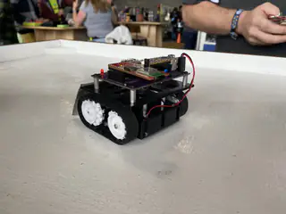 Compact black four-wheeled robot with an exposed circuit board sits alone on a white table.