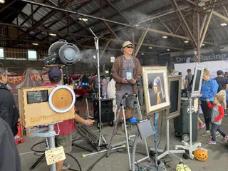 Fog drifts from a round machine above a wooden selfie booth beside framed portraits and an exhibitor.