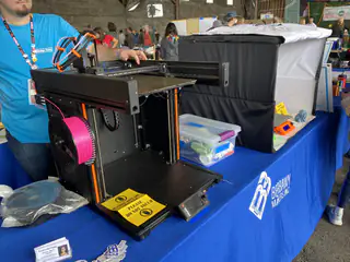 Black open-frame 3D printer with bright pink filament sits on a university display table.