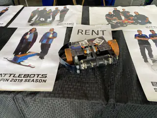 Compact black battle robot with orange side wheels rests on printed team materials.