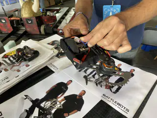 Hands adjust exposed wiring inside a small battle robot above team flyers.