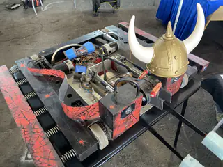 Red-and-black battle robot wears a gold Viking helmet between its exposed drive mechanisms.