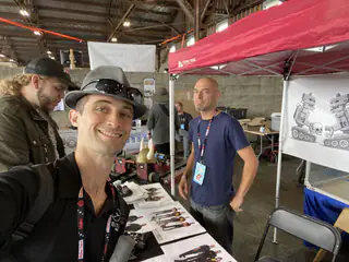 Two smiling exhibitors pose behind a table covered with small robots and team flyers.