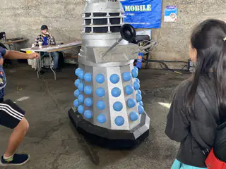 Gray Dalek replica with blue hemispheres stands among visitors inside the exhibition hall.