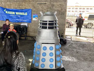 Gray Dalek replica faces a visitor while two seated exhibitors watch from behind.