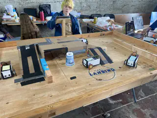 Small wheeled robots occupy a wooden tabletop course marked with a landing zone.