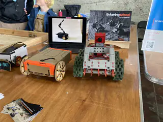 Two wheeled robots stand beside a monitor at a Tinkering School booth.