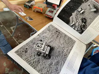 Open book displays black-and-white rover photographs while several small robots sit behind it.