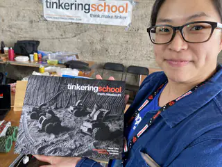 Person wearing glasses holds a Tinkering School project book toward the camera.
