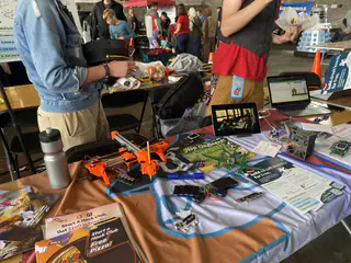 Crowded project table holds an orange quadcopter frame, electronics, monitors, and printed materials.