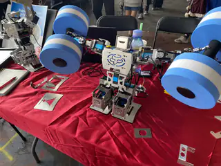 Small white humanoid robot stands before oversized blue foam wheels and scattered electronic parts.
