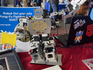 Gray humanoid fighting robot raises one arm beside a laptop and colorful robot props.