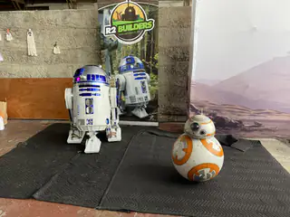 Two R2-D2 replicas and one BB-8 replica stand before an R2 Builders backdrop.