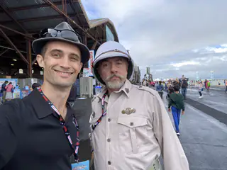 Two attendees pose outdoors, one wearing a beige costume jacket and protective helmet.