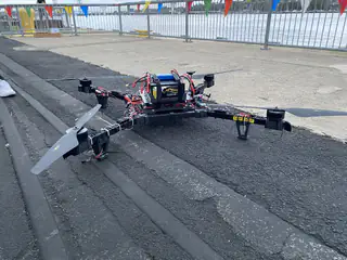 Large four-armed multicopter rests on asphalt with exposed wiring, batteries, and propellers.