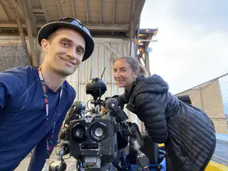 Two smiling adults flank a black humanoid robot whose rounded head faces away.