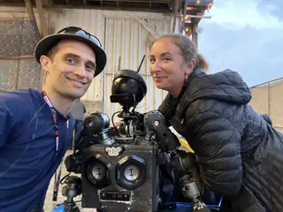 Two adults pose beside the black humanoid robot as its camera-eyed head faces forward.
