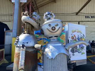 Whimsical silver robot sculpture displayed beside colorful illustrated artwork at Maker Faire.