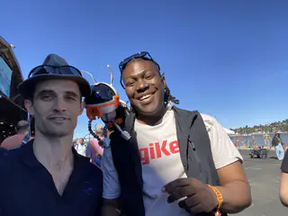 Two smiling exhibitors take a sunny outdoor selfie with costumed attendees behind them.