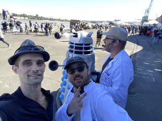Three exhibitors pose with a blue-and-silver Dalek-style robot on the waterfront midway.