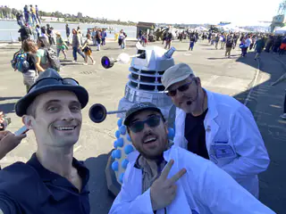 Three smiling exhibitors crowd into a selfie beside the Dalek-style robot and festival visitors.