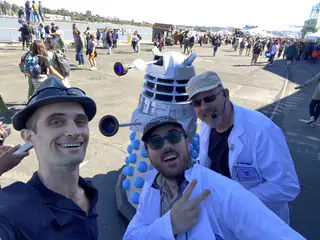 Wide-angle selfie of three exhibitors with the robot and busy outdoor fair behind them.