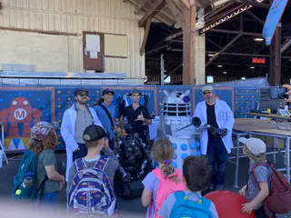 Crowd of young visitors faces the robot demonstration against a colorful Maker Faire backdrop.
