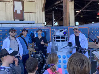 Three presenters speak to children gathered closely around the two display robots.
