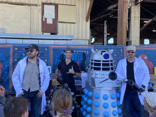 Presenters and a Dalek-style robot face an attentive group of children in the hangar.