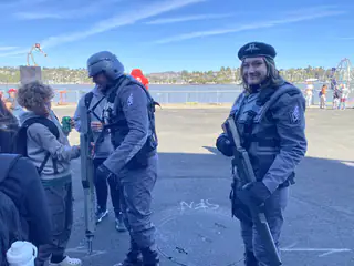 Three costumed attendees in dark utility gear chat on the sunlit waterfront midway.
