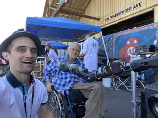 Wide selfie frames two adults and an articulated mechanical arm beside a blue canopy.