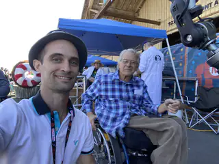 Closer selfie frames two adults beneath a blue canopy, with a mechanical arm cropped above.
