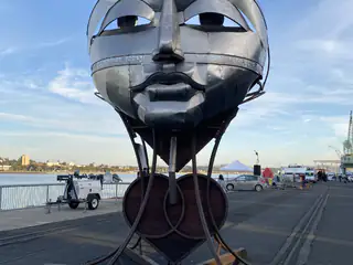 Large sculpted metal face towers above the waterfront with R.O.B. visible in the distance.