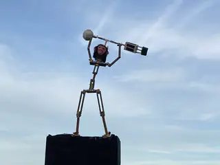 Tall metal sculpture with a monkey head holds a weighted bar against the open sky.