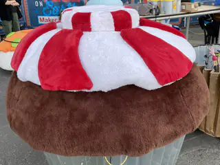 Oversized plush sculpture resembles a brown dome topped with broad red-and-white panels.