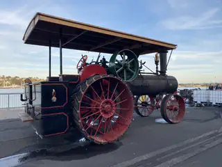 Black-and-red steam tractor with large spoked wheels parked beside the waterfront.