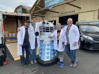 Four exhibitors in white lab coats pose around a blue-and-silver Dalek-style robot.