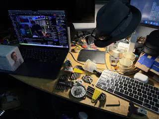 Workbench with an open laptop, keyboard, hand tools, gears, fasteners, and robot parts under a black lamp.