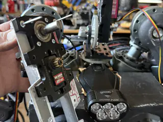 Closer view of the held servo bracket, metal shaft, and small brass gear against the robot.