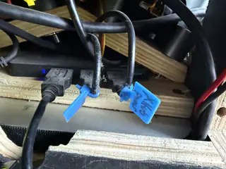 Black cables routed through a wooden frame, each tagged with blue handwritten tape labels.