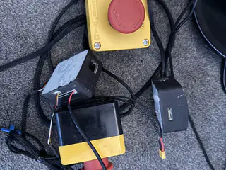 Yellow emergency stop button, battery pack, control boxes, and braided cables arranged on gray carpet.