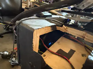 Close view through a rough wooden opening with red and black wires beneath a large gear.
